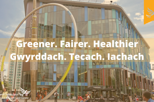 A sunny day looking up at Cardiff Central Library, captioned with the words "Greener. Fairer. Healthier" in both English and Welsh.